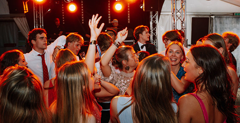 Eine Gruppe junger Erwachsener in festlicher Kleidung tanzt und feiert auf einer Party unter warmen, hellen Scheinwerfern. Viele haben die Hände erhoben, lächeln und lachen in einer ausgelassenen Atmosphäre, während im Hintergrund ein DJ und die Tonanlage zu sehen sind.