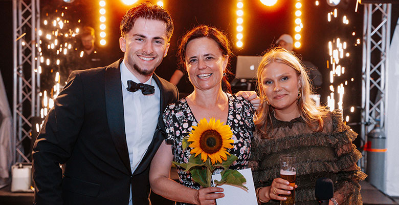 Drei Personen lächeln bei einer Veranstaltung, im Hintergrund sind helle Bühnenlichter und Wunderkerzen zu sehen. Der Mann links trägt einen Smoking, die Frau in der Mitte hält eine Sonnenblume und einen Umschlag in der Hand, und die Frau rechts hält ein Getränk in der Hand und trägt ein Rüschenkleid.