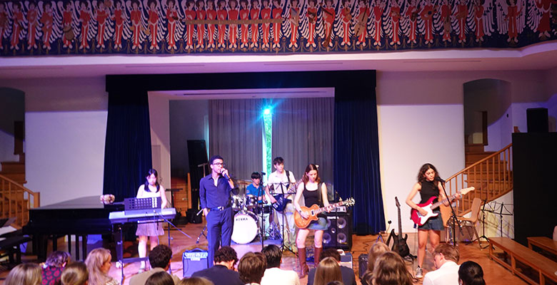 Eine sechsköpfige Band – drei Gitarristen, ein Keyboarder, ein Schlagzeuger und ein Sänger – tritt auf einer Bühne vor Publikum auf. Die Bühne befindet sich in einem Innenraum; links steht ein Klavier, und darüber ist ein dekorativer Fries zu sehen, auf dem mehrere Figuren in Rot und Schwarz dargestellt sind.