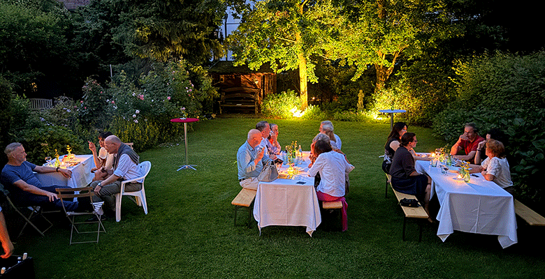 Eine Gruppe von Menschen sitzt in der Abenddämmerung an drei Tischen im Freien auf einer grünen Wiese und genießt das Abendessen. Jeder Tisch ist mit Kerzen beleuchtet und von Bäumen und Gartenpflanzen umgeben. Warmes, sanftes Licht schafft eine gemütliche Atmosphäre für das Beisammensein.