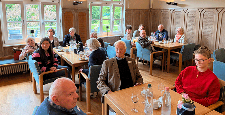 In einem hellen Raum mit großen Fenstern und holzgetäfelten Wänden sitzen eine Gruppe älterer Erwachsener und ein paar jüngere Menschen an Holztischen. Flaschen, Gläser und Blumen stehen auf den Tischen. Die meisten Menschen blicken nach vorne und wirken aufmerksam und engagiert.