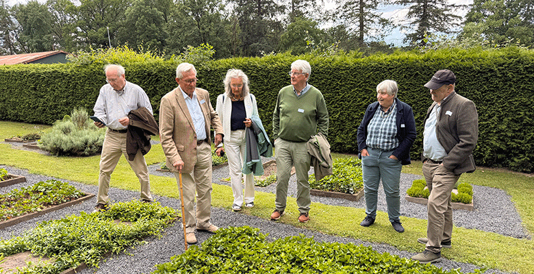 Sechs ältere Erwachsene, vier Männer und zwei Frauen, stehen plaudernd und lächelnd in einem formellen Garten mit gepflegten Hecken und Kieswegen. Sie tragen legere, aber gepflegte Kleidung und haben Jacken in der Hand. Bäume und Grünpflanzen füllen den Hintergrund unter einem bewölkten Himmel.
