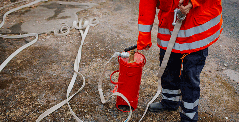 An einem Sommerabend in Soest hält eine Person in einer leuchtend orangefarbenen Warnweste und einer dunklen Hose einen Feuerwehrschlauch in der Hand, der an einen roten Hydranten angeschlossen ist. Mehrere lange, weiße Schläuche liegen draußen auf dem Boden ausgebreitet, wobei nur der Unterkörper zu sehen ist.