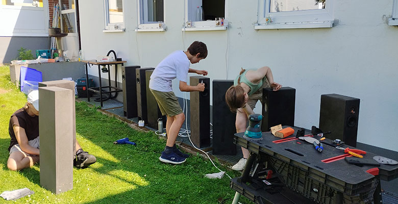An einem sonnigen Tag arbeiten drei Personen im Freien daran, hohe Lautsprecherboxen zusammenzubauen oder zu reparieren. Zwei stehen in der Nähe des Gebäudes und benutzen Werkzeuge, während eine auf dem Rasen sitzt und eine Lautsprecherbox in den Händen hält. Verschiedene Werkzeuge und Ausrüstungsgegenstände liegen verstreut auf einem schwarzen Tisch und auf dem Rasen.