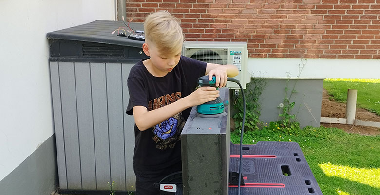 Ein Junge mit blonden Haaren, der ein schwarzes T-Shirt trägt, bearbeitet im Freien eine flache graue Oberfläche mit einem blaugrünen und schwarzen Elektroschleifer. Er steht neben einem Aufbewahrungsschuppen aus Kunststoff und einer Backsteinmauer, im Hintergrund sind Gras und Gartenpflanzen zu sehen.