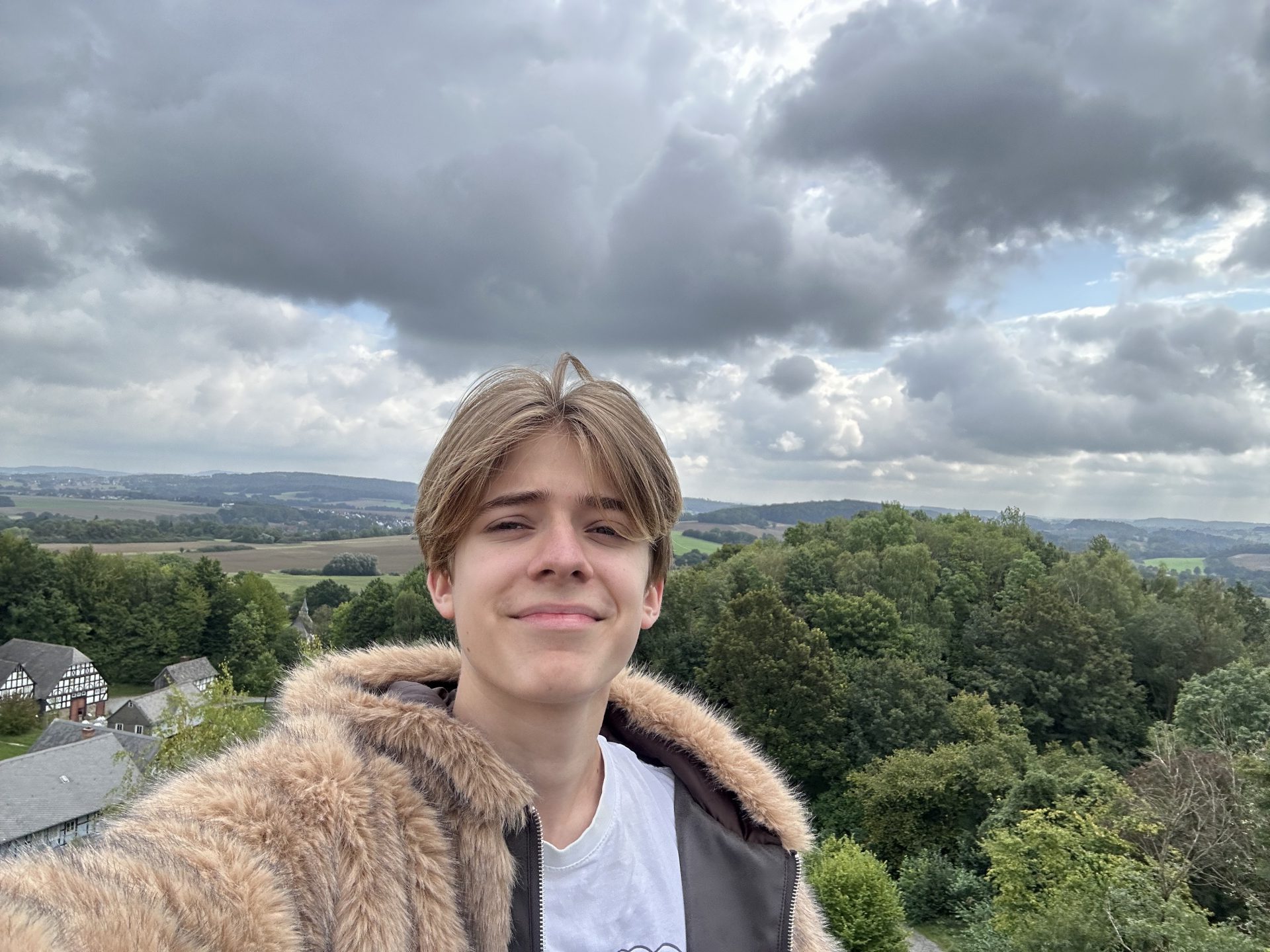 Eine Person mit hellbraunen Haaren, die einen flauschigen, hellbraunen Mantel und ein weißes Hemd trägt, macht draußen ein Selfie. Im Hintergrund sind grüne Bäume, sanfte Hügel, Felder und ein bewölkter Himmel zu sehen. Rechts sind einige Häuser zu erkennen. Das Bild steht auf dem Kopf – Dario Rasch.