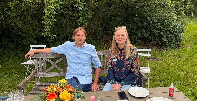 Ein Mann und eine Frau sitzen auf einer Holzbank in einem üppig bewachsenen Garten und genießen einen Sommerabend in Soest. Sie lächeln in die Kamera und sind umgeben von Tellern, Gläsern, einer roten Flasche und leuchtenden Blumen, während grüne Bäume und Gras den Hintergrund bilden.