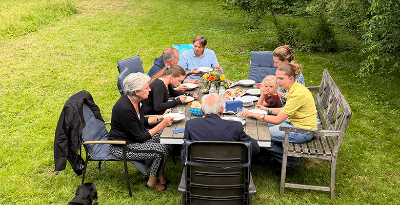 Acht Menschen unterschiedlichen Alters sitzen an einem rechteckigen Tisch im Freien auf dem Rasen und genießen gemeinsam ein Essen an einem Sommerabend in Soest. Teller, Speisen und Getränke stehen auf dem Tisch, während ein schwarzer Hund in der Nähe ruht. Die Gruppe entspannt sich inmitten von Grün und Bäumen.