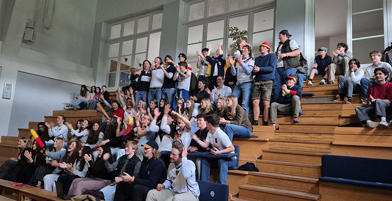 Eine Gruppe von Menschen, überwiegend junge Erwachsene, sitzt und steht auf Holztribünenplätzen in einem hellen Innenraum. Sie jubeln begeistert, schwenken kleine deutsche Flaggen, viele klatschen, lächeln und strecken vor Begeisterung die Arme in die Höhe.