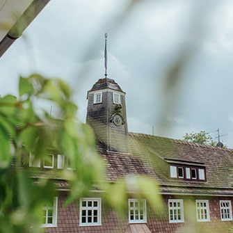 Ein Gebäude mit einem Uhrturm und einer Flagge wird teilweise von grünen Blättern verdeckt. Das Dach ist braun und hat kleine Fenster; der Himmel ist bewölkt. Das Bild ist im Vordergrund aufgrund der Blätter leicht unscharf.