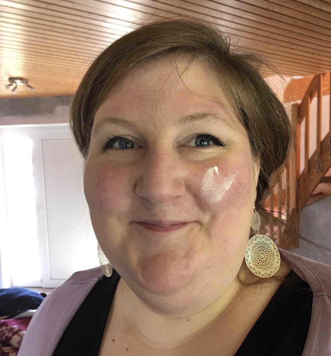 Denise Apel lächelt herzlich, während sie große, runde Spitzenohrringe und ein lila Oberteil trägt. Ein weißer, cremefarbener Streifen ziert ihre linke Wange. Sie befindet sich in einem Innenraum mit Holzdecken und einer Treppe im Hintergrund, während Sonnenlicht durch ein nahegelegenes Fenster oder eine Tür hereinfällt.