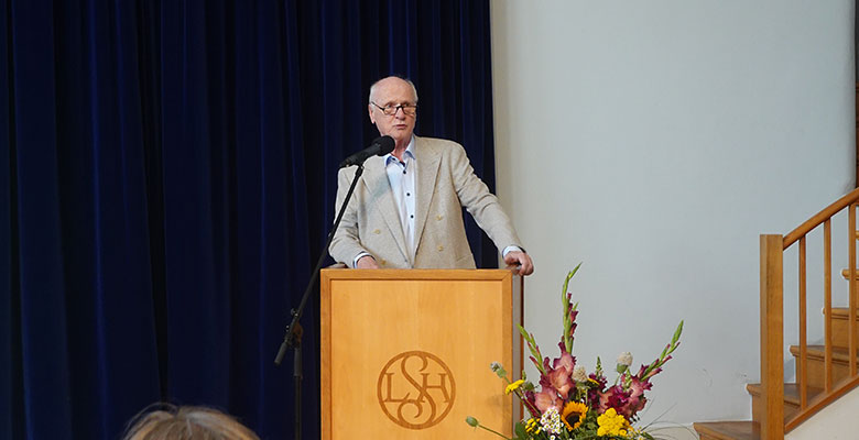 Ein älterer Mann in einem beigen Blazer und mit Brille steht an einem hölzernen Podium, auf dem die Initialen L S H zu sehen sind. Er spricht in ein Mikrofon, rechts von ihm steht ein Blumenarrangement, hinter ihm hängen dunkelblaue Vorhänge. Auf der rechten Seite sind Holztreppen zu sehen.