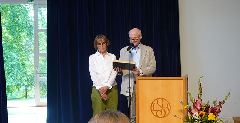 Eine Frau mit Brille und grüner Hose steht neben einem Mann in einer leichten Jacke, der an einem Rednerpult mit Mikrofon von einer Gedenktafel vorliest. Auf dem Rednerpult ist ein LSH-Logo zu sehen. Rechts steht ein Blumenstrauß, und im Hintergrund ist ein blauer Vorhang mit einem Fenster zu sehen.