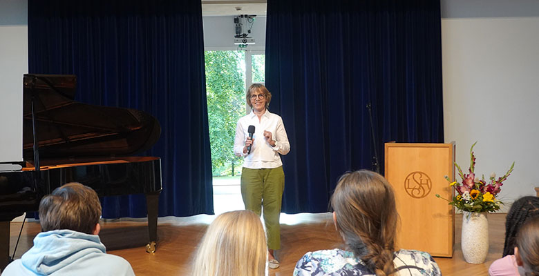 Eine Frau steht mit einem Mikrofon in der Hand auf der Bühne und spricht zu den Zuhörern. Sie trägt eine Brille, ein weißes Hemd und eine grüne Hose. Hinter ihr sind dunkelblaue Vorhänge, ein Flügel, ein Rednerpult mit einem Logo und eine Vase mit Blumen zu sehen. Durch ein Fenster fällt Tageslicht herein.