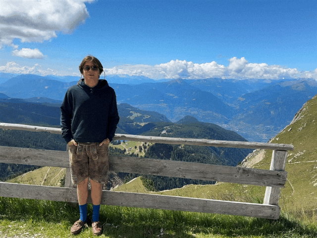 Damian, der eine Sonnenbrille, einen dunklen Kapuzenpulli, braune Shorts und blaue Socken trägt, steht vor einem Holzzaun auf einem grasbewachsenen Hügel und genießt den Panoramablick auf Berge, Täler und einen blauen, mit Wolken übersäten Himmel.