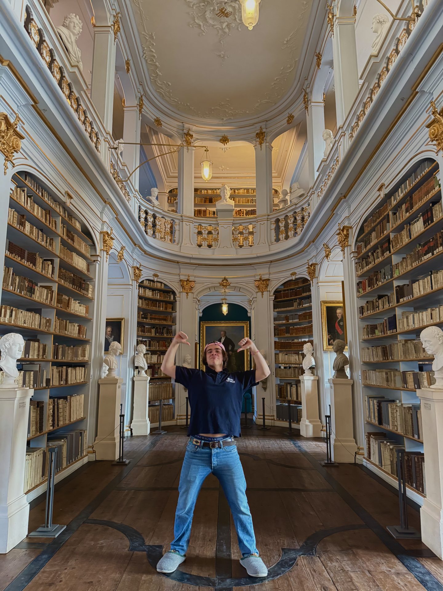 Max-Henry, gekleidet in Jeans, Hausschuhe und ein schwarzes Hemd, steht spielerisch in einer prächtigen, reich verzierten Bibliothek mit hohen Regalen, gewölbten Balkonen, goldenen Akzenten und Marmorbüsten. Er zeigt nach oben, während im Hintergrund Bücher und Kronleuchter glänzen.