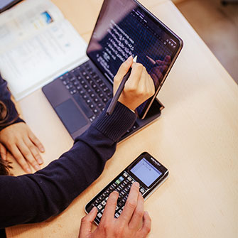Eine Person schreibt mit einem Stylus auf den Bildschirm eines Tablets, während sie mit der anderen Hand Tasten eines Grafikrechners drückt. Auf dem Schreibtisch sind außerdem ein Blatt Papier und ein Laptop zu sehen. Die Szene vermittelt den Eindruck, dass hier gelernt, gearbeitet oder mit Hilfe mehrerer Geräte mathematische Aufgaben gelöst werden.