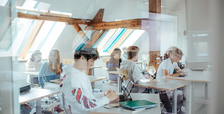 Eine Gruppe von Schülern sitzt an Schreibtischen in einem hellen Klassenzimmer mit großen Fenstern und Holzbalken. Einige nutzen Laptops, während andere sich unterhalten oder schreiben. Sonnenlicht strömt herein und schafft eine ruhige und konzentrierte Atmosphäre. Die Szene wird durch eine Glasscheibe betrachtet.