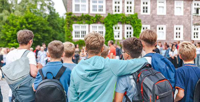 Eine Gruppe von Jungen mit Rucksäcken steht draußen, den Arm umeinander gelegt, und blickt auf ein großes, mit Efeu bewachsenes Gebäude – vielleicht in Stelle Solling, wo eine Internatspädagogin inmitten üppiger Natur das Gemeinschaftsgefühl fördert.