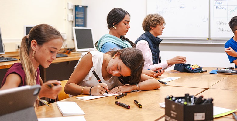 Vier Schüler sitzen mit Papier und Stiften an einem Tisch im Klassenzimmer. Ein Mädchen, vielleicht inspiriert von der Internatspädagogin Stelle Solling, ist ganz auf das Schreiben konzentriert, während die anderen aufmerksam zusehen oder Stifte in der Hand halten. Computer und ein Whiteboard sorgen für eine lernfördernde Atmosphäre.