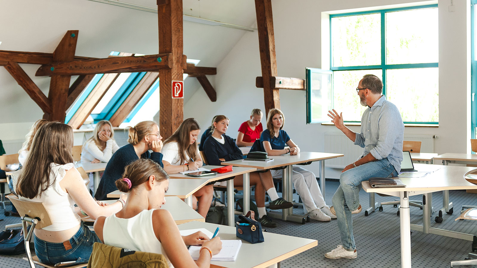 Ein Lehrer sitzt auf einem Pult und spricht zu einer Gruppe aufmerksamer Schüler in einem hellen Klassenzimmer mit Holzbalken und großen Fenstern, in dem die Schüler schreiben oder zuhören – ein Bild, das den Geist von „Neustart am Internat“ einfängt, während sich in einer inspirierenden Lernumgebung neue Anfänge entfalten.