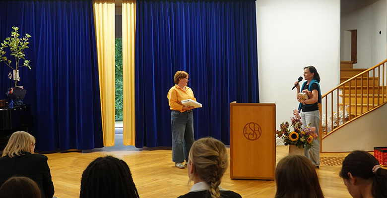 Eine Frau in einem orangefarbenen Shirt steht auf der Bühne, hält Bücher in der Hand und blickt auf eine Frau mit einem Mikrofon hinter einem Rednerpult mit einem Blumenlogo. Im Vordergrund sitzen Zuschauer. Im Hintergrund sind blaue Vorhänge und eine Treppe zu sehen. Auf der Bühne stehen ein Blumenstrauß und ein Topfbaum.