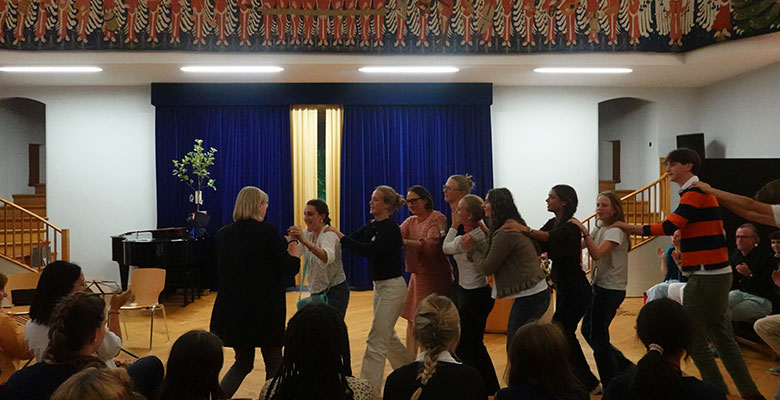 Eine Gruppe von Menschen bildet eine Conga-Reihe und tanzt in einem hell erleuchteten Saal mit einem blauen Vorhang als Hintergrund, Holzböden und einem Publikum, das davor sitzt. Links stehen ein Klavier und eine Zierpflanze, und bunte Muster zieren den oberen Teil der Wand.