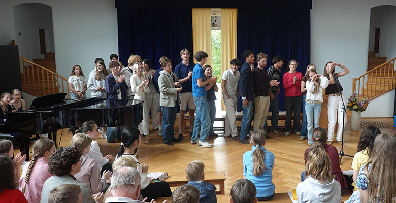 Eine Gruppe von Schülern steht auf einer Holzbühne vor einem Publikum; einige klatschen oder lächeln. Das Publikum sitzt auf Bänken und klatscht ebenfalls. Auf der Bühne stehen ein Klavier und ein Blumenstrauß, im Hintergrund sind blaue Vorhänge und ein helles Fenster zu sehen.