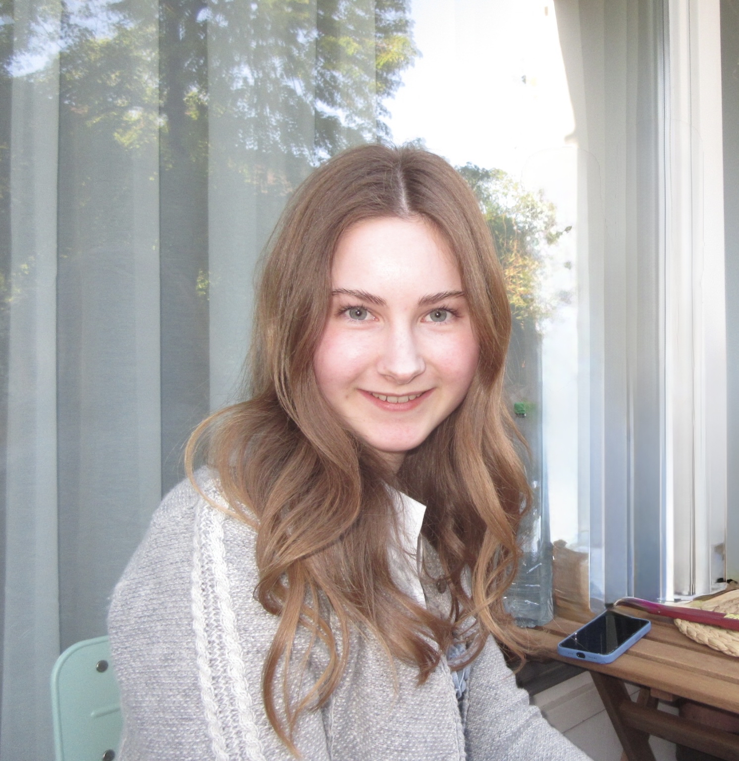 Eine junge Frau mit langen, welligen braunen Haaren und blauen Augen lächelt in die Kamera. Sie trägt einen hellgrauen Pullover und sitzt draußen vor einem Fenster mit transparenten Vorhängen. Auf dem Holztisch neben ihr liegen ein Handy und ein Buch – ein spontaner Moment aus Catharina Frehoffs Alltag.