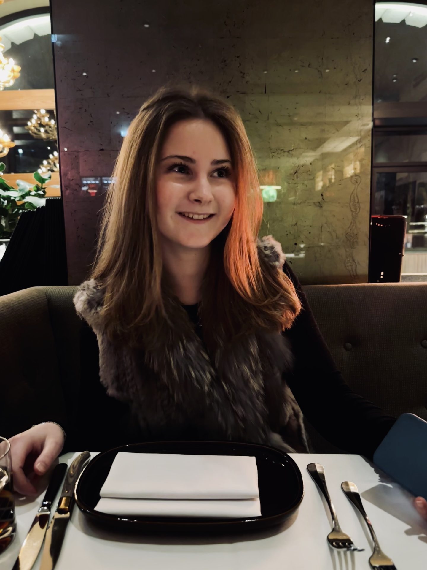 Catharina Frehoff, eine Frau mit langen braunen Haaren, sitzt an einem Restauranttisch, der mit schwarzen Tellern und Besteck gedeckt ist. Sie trägt eine pelzige Weste über einem dunklen Oberteil und lächelt, während sie zur Seite blickt. Der Hintergrund ist warm beleuchtet, und man sieht Reflexionen sowie dekorative Lichter.