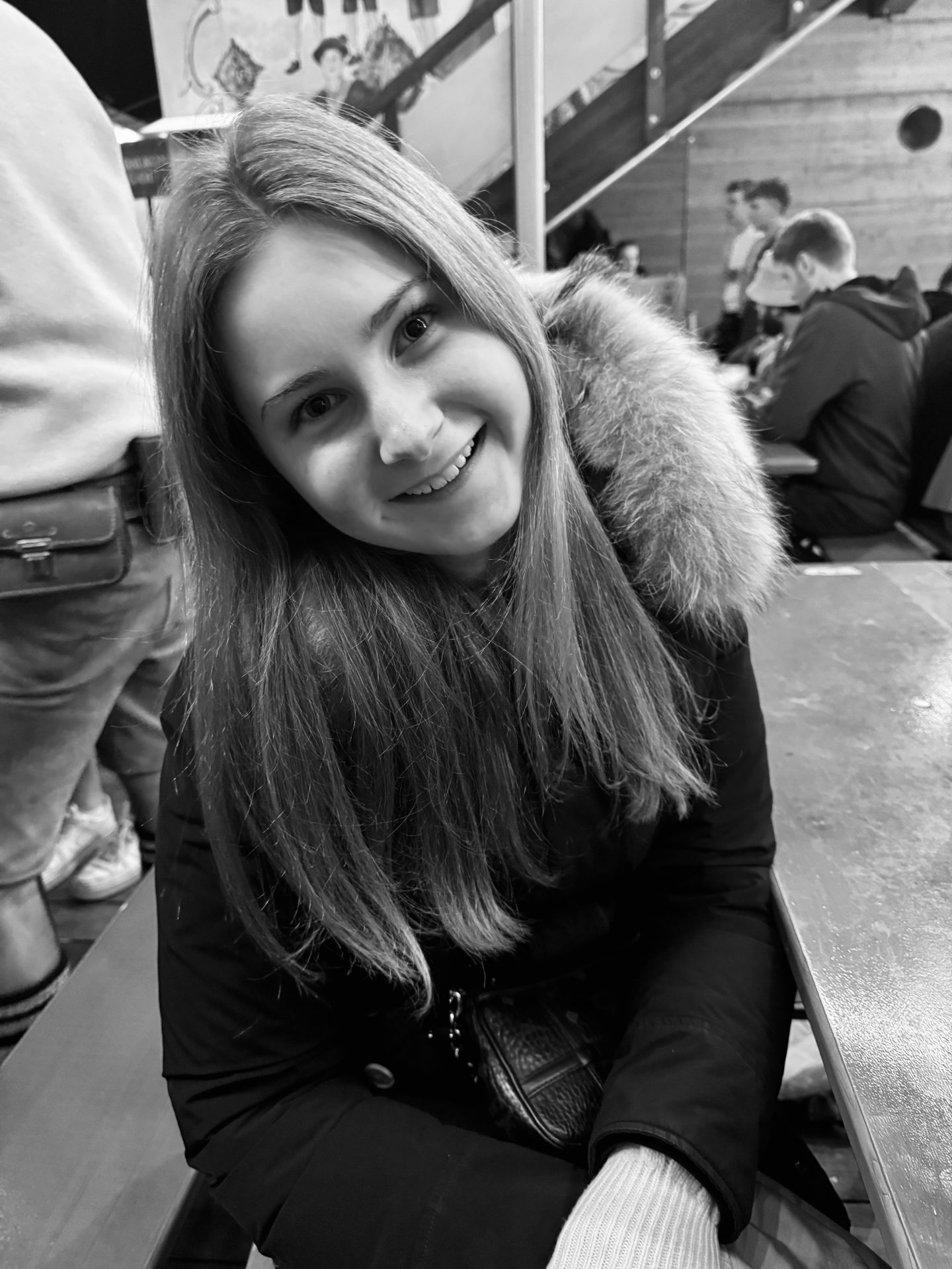 Eine junge Frau mit langen Haaren und einem Wintermantel mit pelzbesetzter Kapuze sitzt auf einer Bank in einem Innenraum und lächelt. Catharina Frehoff befindet sich unter den sitzenden und stehenden Personen im Hintergrund. Das Bild ist in Schwarz-Weiß gehalten.