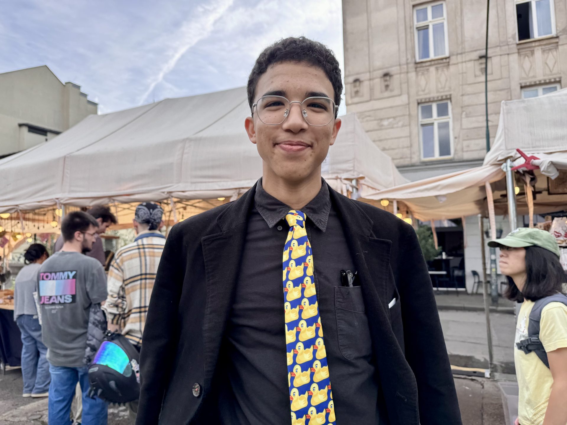 Max–Henry, ein junger Mensch mit Brille und lockigem Haar, lächelt auf einem Markt im Freien. Er trägt einen schwarzen Anzug, ein schwarzes Hemd und eine Krawatte mit gelben Gummienten. Im Hintergrund sind Menschen und Verkaufszelte zu sehen. Die Atmosphäre wirkt ungezwungen und lebhaft.