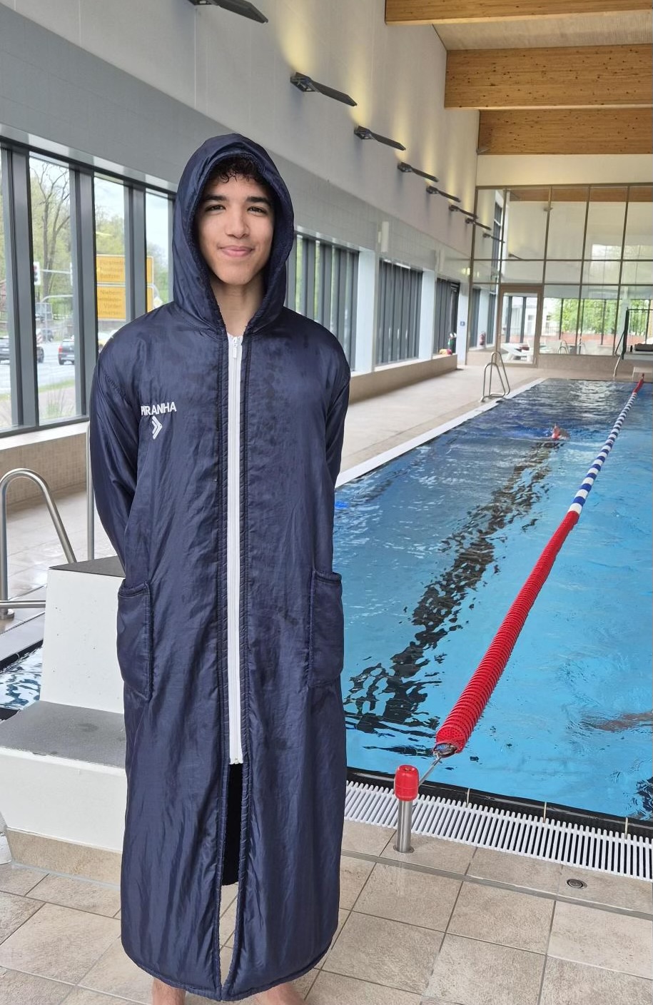 Ein lächelnder Teenager namens Max–Henry steht in einem dunkelblauen Schwimmparka am Rand eines Hallenbads. Die Kapuze ist hochgezogen, und der Parka ist bis oben zugeknöpft. Hinter ihm befindet sich ein helles Schwimmbecken, in dem eine Person in einer Bahn schwimmt. Große Fenster lassen Tageslicht herein. Der Beckenrand ist gefliest und größtenteils leer.