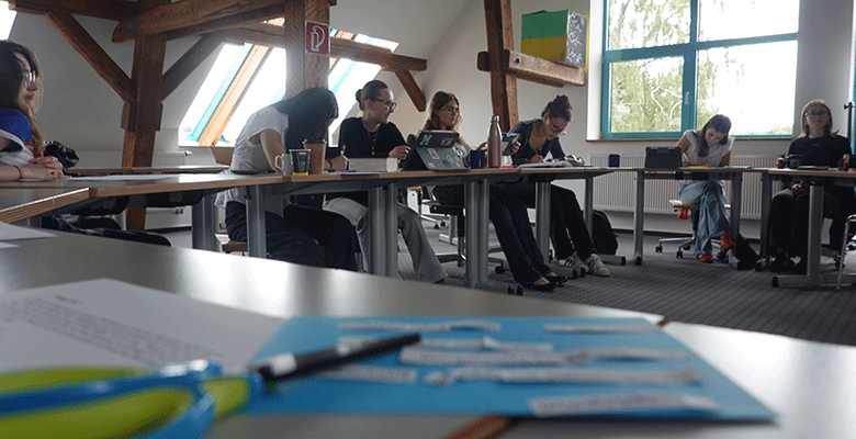 Eine Gruppe von Menschen sitzt an einem U-förmigen Tisch in einem hellen Raum mit Holzbalken und ist ganz auf das Schreiben oder die Diskussion konzentriert. Auf den Tischen liegen Papiere, Stifte und Getränke. Im Vordergrund sind verschwommen Papier, ein Stift und eine Schere zu sehen. Durch ein großes Fenster fällt Tageslicht herein.