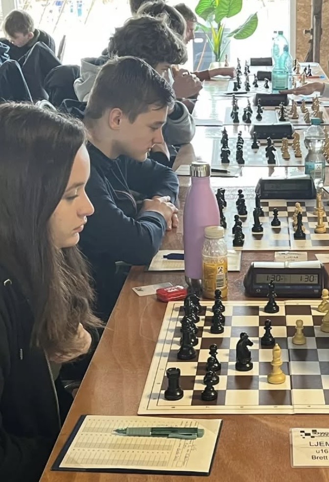 Mehrere junge Leute sitzen an einem langen Tisch und spielen Schachpartien. Anna (LSH seit 2023) mit langen braunen Haaren konzentriert sich auf ihre Partie; neben ihr liegen ein Notizblatt und eine Trinkflasche. Weitere Spieler und digitale Schachuhren füllen den hellen Raum.