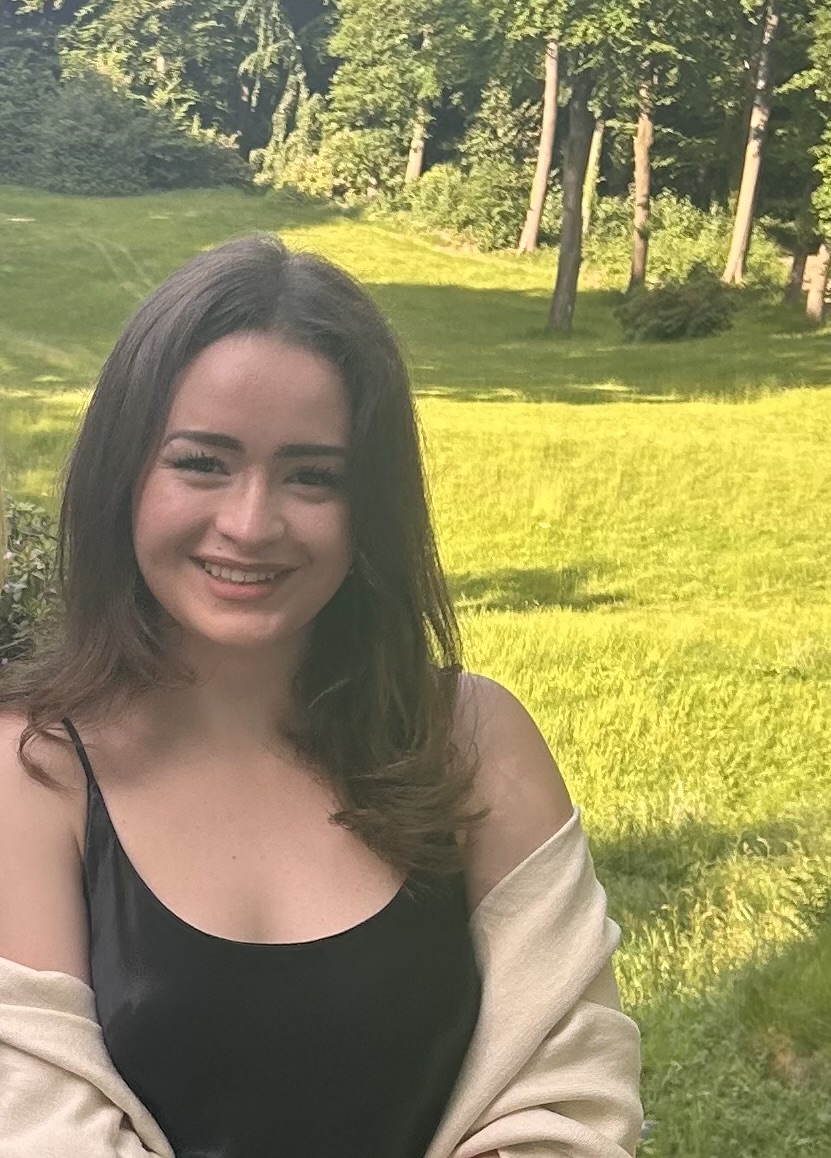 Anna (LSH seit 2023), eine junge Frau mit langen braunen Haaren, die ein schwarzes, ärmelloses Kleid und einen beigen Schal trägt, lächelt in die Kamera. Sie steht auf einer sonnenbeschienenen grünen Wiese, im Hintergrund sind hohe Bäume und dichtes Laubwerk zu sehen.