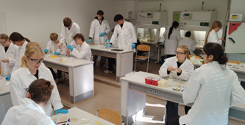 Studierende in weißen Laborkitteln und mit Schutzbrillen arbeiten in einem geschäftigen Labor und hantieren an ihren Arbeitsplätzen mit Petrischalen und Laborgeräten. Einige benutzen Pipetten, während andere Ergebnisse besprechen. Im Hintergrund sind Geräte und Abzüge zu sehen.