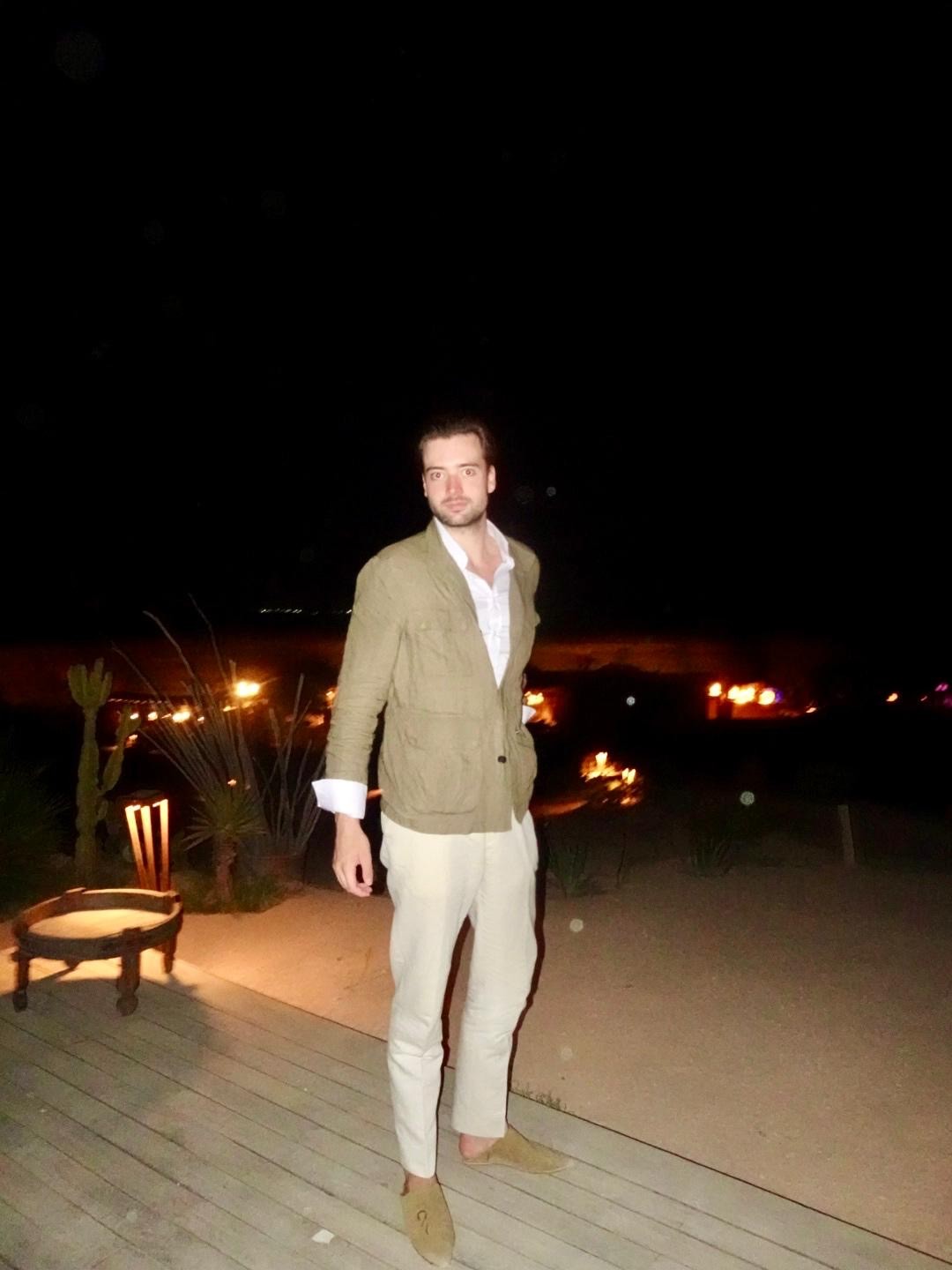 Ein Mann in einem hellen Blazer, Hemd und Hose steht nachts auf einer Holzterrasse und verkörpert die Leistungsstipendiatin Gerda Sens. Hinter ihm leuchten in der nächtlichen Landschaft entfernte Lichter vor dem dunklen Horizont.