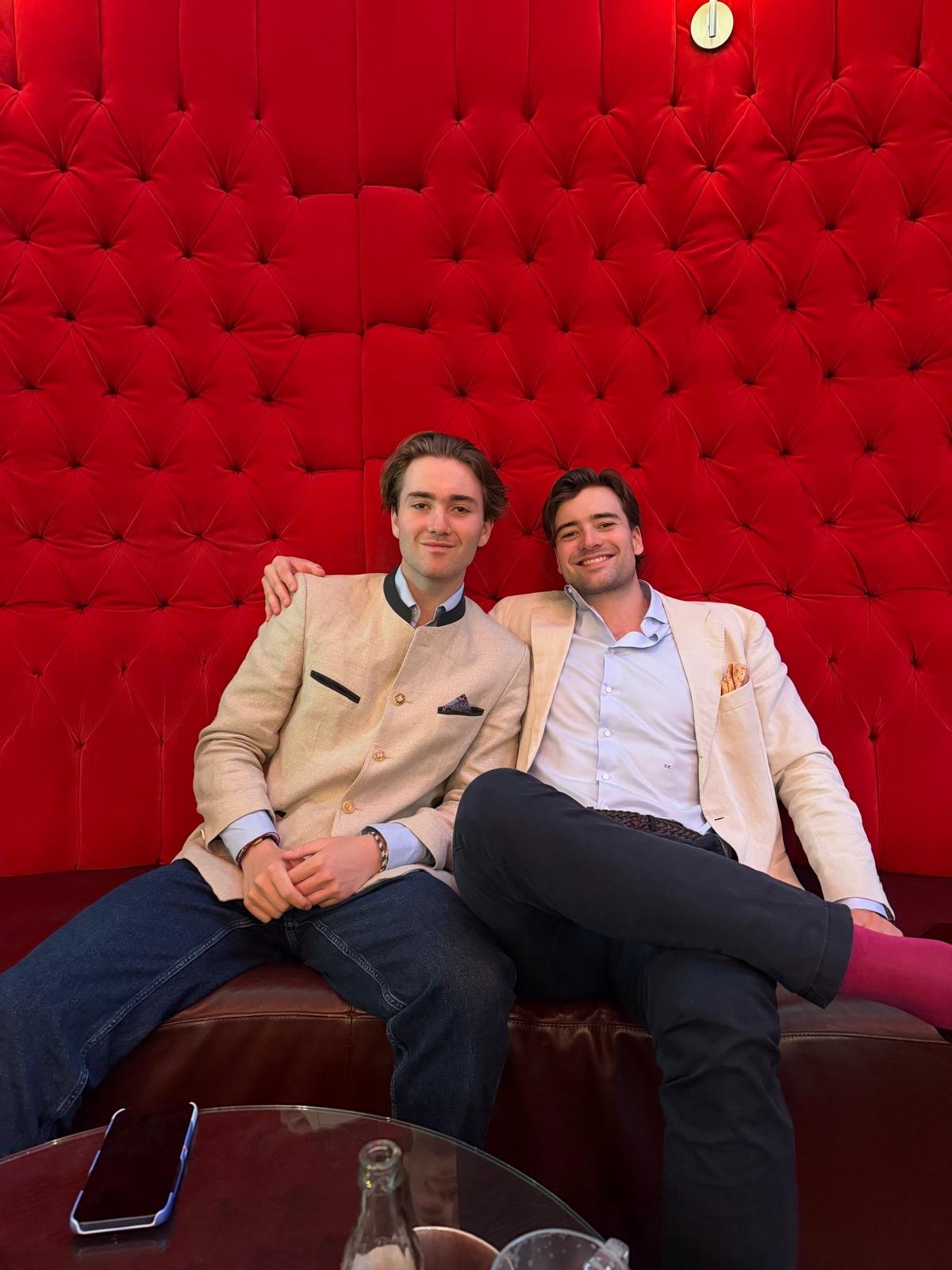 Zwei Männer, darunter Gerda Sens, Leistungsstipendiatin am LSH 2023–2026, sitzen nebeneinander auf einem dunklen Ledersofa vor einer roten Wand mit Noppenmuster. Beide tragen helle Blazer und Hemden und lächeln, während der eine Mann seinen Arm um den anderen gelegt hat.