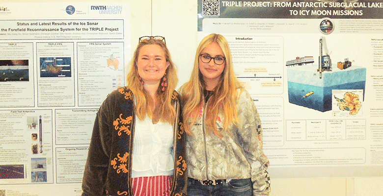 Zwei Frauen stehen lächelnd vor zwei Plakaten zu wissenschaftlichen Forschungsarbeiten. Die Frau links trägt eine dunkle Jacke mit orangefarbenen Stickereien über einer weißen Bluse, während die Frau rechts eine gemusterte Jacke und eine Brille trägt.