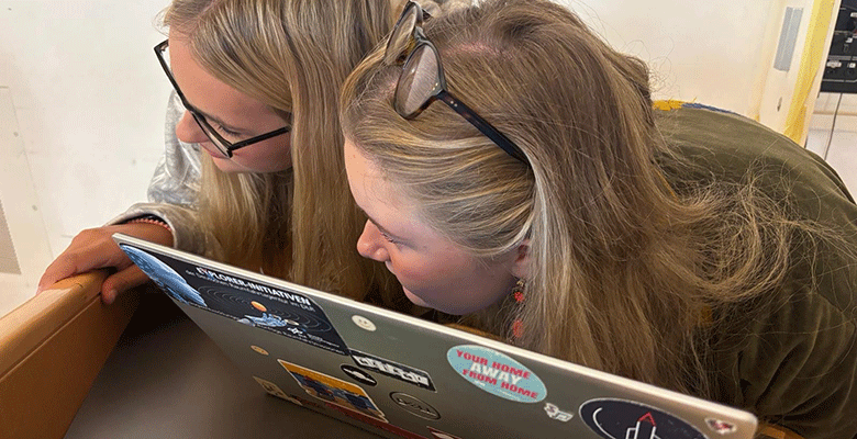 Zwei junge Frauen mit langen blonden Haaren beugen sich über einen Laptop und blicken konzentriert auf den Bildschirm. Der Deckel des Laptops ist mit verschiedenen Aufklebern beklebt, darunter einer mit der Aufschrift Your home away from home.