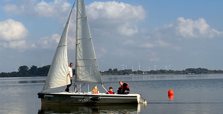 Ein Segelboot mit vier Personen an Bord gleitet über einen ruhigen See in der Nähe von Labor und Moor. Unter einem blauen Himmel mit vereinzelten Wolken sind entlang des entfernten Ufers Windkraftanlagen zu sehen.
