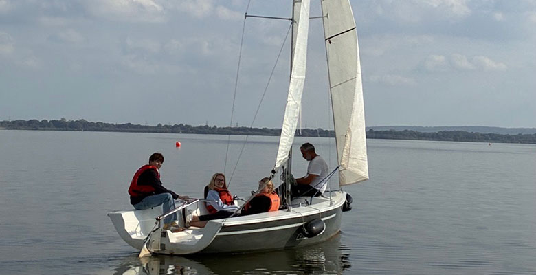 Drei Personen, die Schwimmwesten tragen, sitzen in einem kleinen Segelboot auf einem ruhigen See. Eine Person sitzt am Heck und steuert das Boot, während zwei weitere im Cockpit unter einem weißen Segel sitzen.