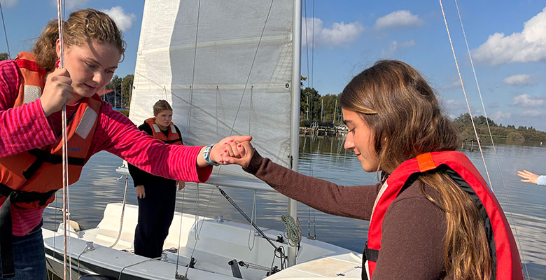 Zwei junge Menschen auf einem Segelboot reichen sich über das Deck hinweg die Hände. Eine dritte Person steht im Hintergrund auf dem Boot.